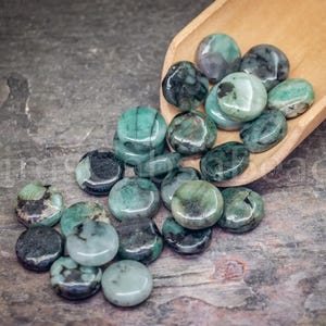 May include: A collection of polished emerald green gemstones, some with dark inclusions, spilling from a small wooden scoop onto a textured gray surface. The gemstones vary in shade, from light to deep green.