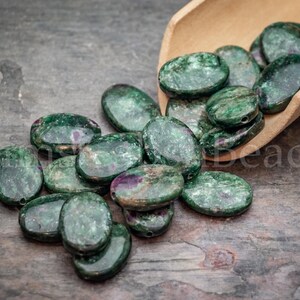 May include: Oval-shaped green beads with red and purple inclusions are scattered on a wooden surface, some spilling from a wooden scoop. The beads have a polished, glossy finish, and are suitable for jewelry making.