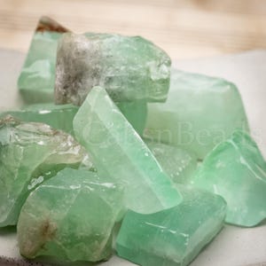 May include: A collection of raw, uncut green calcite crystals. The crystals vary in size and shape, with a translucent, glassy appearance. Some pieces have a lighter, almost white, hue.