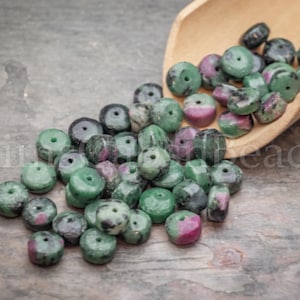 May include: A collection of green and pink gemstone beads with a hole drilled through the center. The beads are spilling out of a small wooden scoop. The beads have a mix of green and pink colors and are cylindrical in shape.