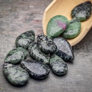 May include: A collection of teardrop-shaped gemstones in shades of green and black, some with a pinkish-red center. The stones are scattered on a gray surface, with a wooden scoop holding a few more. The gemstones are likely for jewelry making.