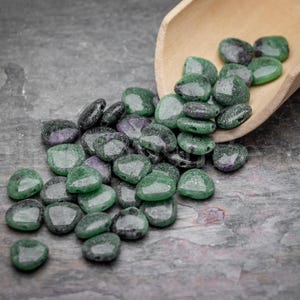 May include: A collection of green and purple glass beads, some with a faceted design, spilling from a wooden scoop onto a textured gray surface. The beads vary in size and shade, creating a visually appealing display.