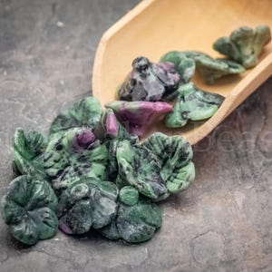May include: A collection of carved gemstone beads in the shape of flowers, with green and purple hues. The beads are scattered on a gray surface, with some spilling from a wooden scoop.