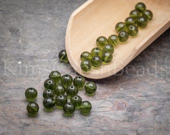 Può includere: Una collezione di piccole perline rotonde e traslucide di colore verde oliva. Alcune perline sono raggruppate su una superficie piana, mentre altre sono contenute in una paletta di legno chiaro. Le perline presentano una finitura liscia e lucida.