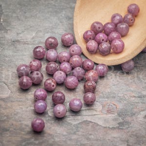 May include: A collection of round, faceted, dusty rose-colored gemstone beads scattered across a surface, with several beads resting inside a light-colored wooden scoop. The beads exhibit a semi-translucent quality with natural inclusions.
