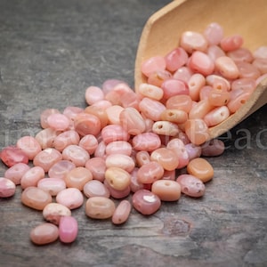 May include: A collection of pink gemstone beads, tumbled and polished, spilling from a small wooden scoop onto a textured gray surface. The beads vary in shade from pale peach to deeper rose, with visible holes for stringing. The text "King Beads" is visible.