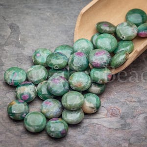 May include: A collection of green and pink gemstone beads, some with visible holes, are scattered on a textured surface. A wooden scoop holds additional beads. The beads have a polished, rounded shape, and the word "beads" is partially visible.
