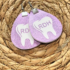 May include: Pair of lavender teardrop earrings with a white tooth graphic and the letters "RD" printed on each. The earrings have silver-colored hooks and are displayed on a woven background.