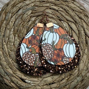 Puede incluir: Pendientes en forma de lágrima con temática otoñal. La capa superior presenta un estampado de calabazas en naranja y blanco, mientras que la capa inferior es marrón brillante. Los pendientes cuelgan de ganchos dorados.