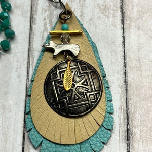 May include: A layered pendant necklace with a turquoise, tan, and bronze color scheme. The pendant features a teardrop shape with fringe, a metal charm, and a decorative circular element. The necklace has turquoise beads.