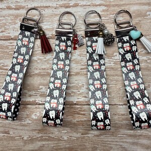 May include: Four wristlet keychains featuring a black background with a repeating pattern of cartoon teeth wearing red scarves. Each keychain has a silver metal clasp, a split ring, and a unique charm including tassels, a heart, or beads.
