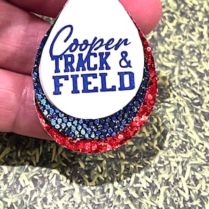 May include: A teardrop-shaped earring with the text "Cooper TRACK & FIELD" in blue. The earring has layers of red, blue, and white glitter. A silver loop is at the top for hanging.