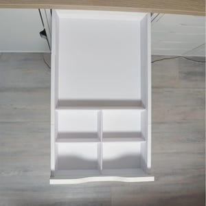 May include: A white, open drawer with a divided interior. The drawer is rectangular with a curved bottom edge and is divided into four smaller compartments. The drawer is set against a light wood-toned surface.