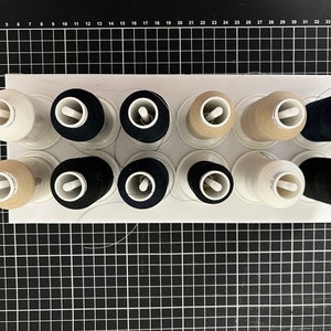 May include: A white plastic thread organiser with 10 spools of thread. The spools are in three colours: white, beige, and black.