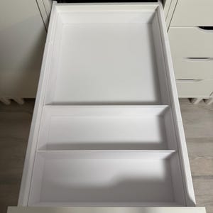 Könnte beinhalten: Eine weiße Schublade, die vollständig ausgezogen ist und eine glatte Innenfläche aufweist. Die Schublade ist durch zwei horizontale weiße Trennwände in drei separate Bereiche unterteilt, die für Ordnung und Aufbewahrung sorgen.