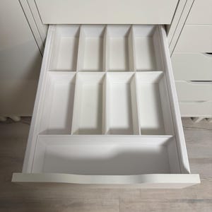 Puede incluir: Cajón de cocina blanco abierto con un organizador de cubiertos incorporado. El cajón está dividido en múltiples compartimentos para guardar utensilios. El cajón es parte de un gabinete blanco con una encimera de madera clara.