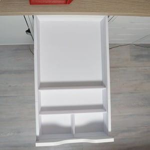 May include: A white, open drawer with multiple compartments and shelves. The drawer is pulled out from a light-colored wooden desk. The interior of the drawer is divided into sections, offering storage solutions. The drawer is empty.