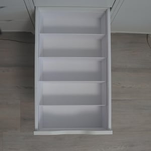 May include: An open white drawer with four evenly spaced shelves. The drawer is part of a larger white cabinet, with a view from above. The drawer is empty and ready for organization. The surrounding floor is a light wood grain.