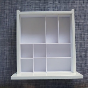 Op de afbeelding: Een witte displayplank met een raamachtig ontwerp, met meerdere compartimenten van verschillende afmetingen. De plank is verdeeld in rechthoekige en vierkante secties, ideaal voor het organiseren van kleine voorwerpen. De plank is gemaakt van hout en heeft een platte basis.