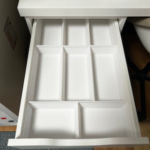 Peut inclure: Un tiroir de bureau blanc avec plusieurs compartiments, idéal pour organiser de petits objets. Le tiroir est ouvert, révélant les séparateurs intérieurs. Le bureau est blanc, et le tiroir est sorti.