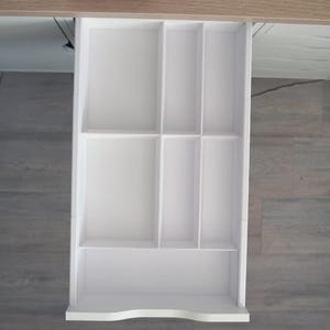 May include: A white wooden drawer organizer with multiple compartments of varying sizes. The organizer is designed to fit inside a drawer and has a curved bottom edge. The drawer is open, revealing the interior organization.