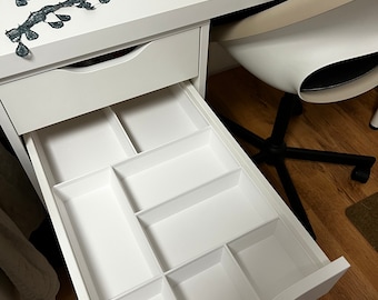 May include: White desk with an open drawer featuring a divided organizer. The drawer is sectioned into multiple compartments of varying sizes. Dark teal trim is visible on the desk surface.