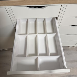 Può includere: Un cassetto bianco con un organizer aperto e diviso. L'organizer ha più scomparti rettangolari di varie dimensioni. Il cassetto fa parte di un mobile bianco con un piano di lavoro color legno chiaro. Un'etichetta nera con la parola "Rotary" è visibile.