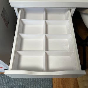 Op de afbeelding: Een open witte bureaulade, verdeeld in acht rechthoekige compartimenten. De binnenkant van de lade is zichtbaar en toont de verdelers. Het bureau heeft een modern design met een gladde, witte afwerking. De lade staat tegen een neutrale achtergrond.