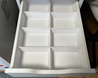 May include: A white desk drawer with eight rectangular compartments. The drawer is open, revealing the interior dividers. The desk has a clean, modern design, with a smooth, white finish. The drawer is set against a neutral background.