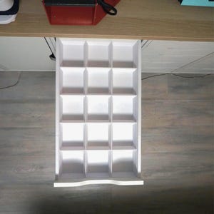 May include: A white, divided drawer organizer with sixteen square compartments. The organizer is pulled out from a light-colored wooden cabinet. The drawer is designed to hold small items, providing organized storage.