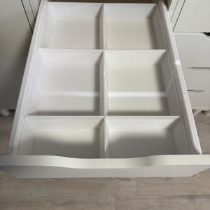May include: A white drawer with six compartments, open and ready for organization. The drawer is part of a larger white cabinet, with a clean and modern design. The compartments are evenly sized, offering versatile storage options.