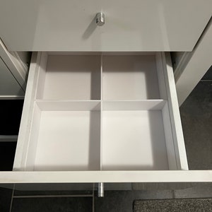 May include: A white drawer with a silver handle is open, revealing a white drawer organizer with four compartments.