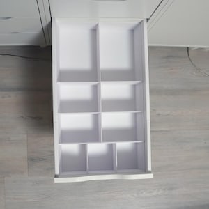 Op de afbeelding: Een open witte lade met meerdere compartimenten voor opslag. De lade is verdeeld in verschillende secties door verticale en horizontale verdelers. De lade maakt deel uit van een grotere witte kast en is ontworpen voor het organiseren van kleine items.