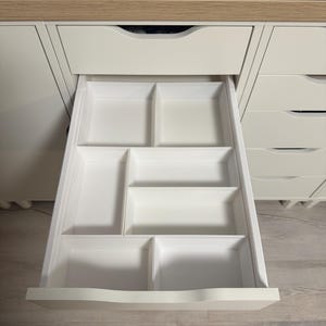 May include: Open white drawer with multiple compartments for organizing items. The drawer is part of a white cabinet with a light wood-colored countertop. A black rectangular object sits on the countertop.