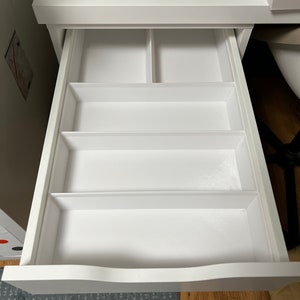 Könnte beinhalten: Geöffnete weiße Schreibtischschublade mit mehreren Fächern zur Aufbewahrung. Die Schublade bietet rechteckige und quadratische Abschnitte, ideal zum Organisieren kleiner Gegenstände. Die Schublade ist aus weißem Holz gefertigt und hat eine geschwungene Vorderkante.