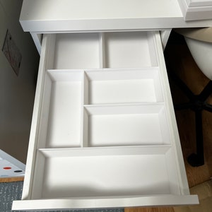 Puede incluir: Cajón de escritorio blanco, abierto y vacío, con varios compartimentos integrados de diferentes tamaños. El cajón está limpio y parece estar hecho de madera o un material similar. La superficie del escritorio sobre el cajón también es blanca, con carretes de hilo visibles.