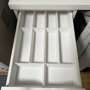 Può includere: Un cassetto da scrivania bianco aperto con più scomparti rettangolari. Il cassetto è diviso in varie sezioni, creando uno spazio di archiviazione organizzato. I compartimenti sono progettati per contenere piccoli oggetti.