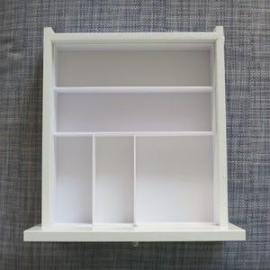 May include: A white wooden organizer with multiple compartments and shelves. The organizer has a rectangular shape with a flat base and a top shelf. The interior is divided into various sections, providing storage space for small items. The overall design is clean and minimalist.