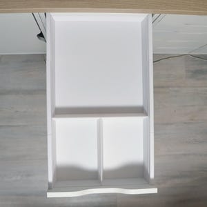 Op de afbeelding: Een witte rechthoekige lade die uit een bureau van licht hout is getrokken. De binnenkant van de lade is verdeeld in drie aparte secties, met één groot rechthoekig vak aan de bovenkant en twee kleinere vierkante vakken aan de onderkant.