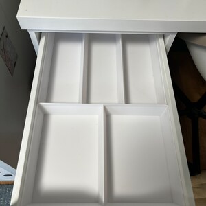 Op de afbeelding: Een witte bureau met een open lade, met een verdeeld interieur. De lade is verdeeld in compartimenten, ideaal voor het organiseren van kleine items. Naaibenodigdheden zijn zichtbaar op het bureaublad boven de lade.