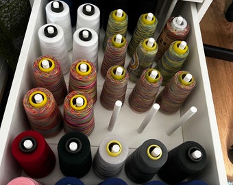 May include: A drawer filled with spools of thread in various colors, including white, red, pink, blue, green, gray, and black. The spools are organized in rows and columns.
