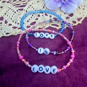 May include: Three beaded bracelets with the words "LOVE IS LOVE" spelled out in white letters. The bracelets are layered on a purple fabric background with a purple and white flower.