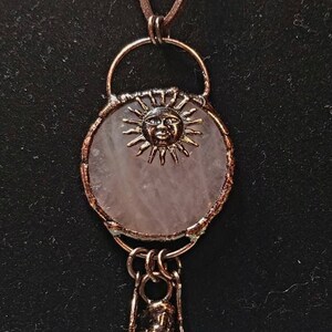 Op de afbeelding: Een ketting met een hanger van rozenkwarts, een koperen zonhanger en een punt van helder kwartskristal met een blauwe steenaanvulling. De ketting is aan een bruin leren koord.