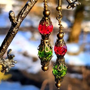 May include: A pair of dangle earrings with green and red glass beads. The earrings have a gold-tone metal design and are hanging from a branch.