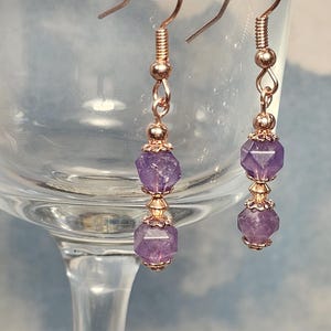 May include: A pair of dangle earrings featuring faceted purple gemstones and rose gold-colored metal accents. The earrings have a delicate design with a hook closure and are suspended from a series of small beads and decorative caps.