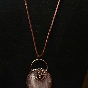 Op de afbeelding: Een roze rozenkwarts hangerketting met een zonontwerp en een hanger van helder kwartskristal. De ketting is gemaakt van bruin lederen koord en blauwe lapis lazuli kralen. Passende oorbellen zijn ook inbegrepen.