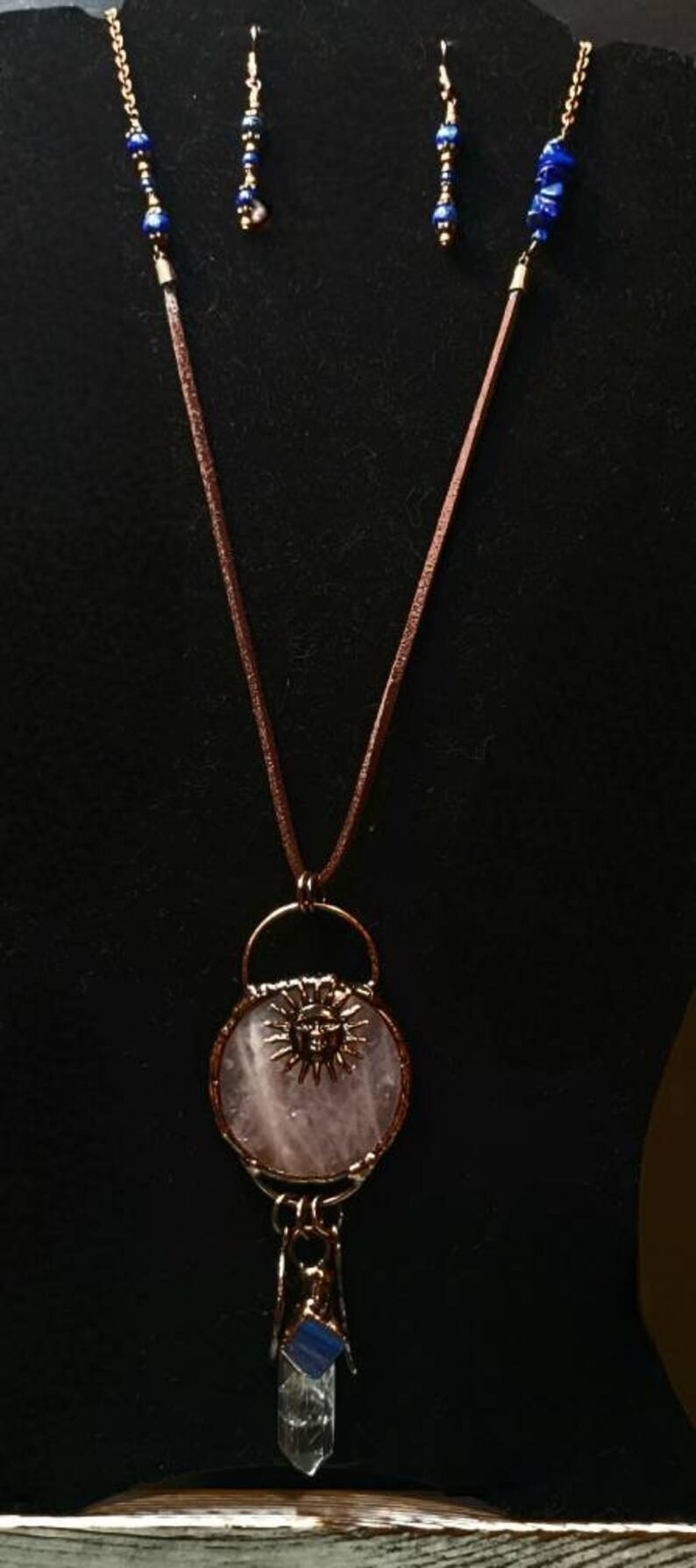 Op de afbeelding: Een roze rozenkwarts hangerketting met een zonontwerp en een hanger van helder kwartskristal. De ketting is gemaakt van bruin lederen koord en blauwe lapis lazuli kralen. Passende oorbellen zijn ook inbegrepen.