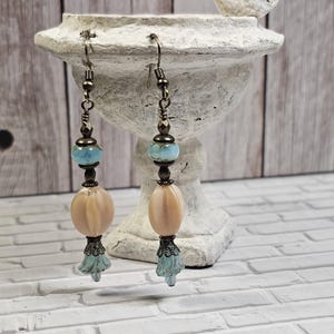 May include: A pair of dangle earrings with a light blue and peach colored bead, a bronze colored bead, and a light blue flower-shaped charm.