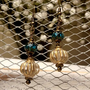 May include: A pair of dangle earrings with a gold tone finish. The earrings feature a clear glass bead with a gold metallic finish and a teal blue faceted glass bead.