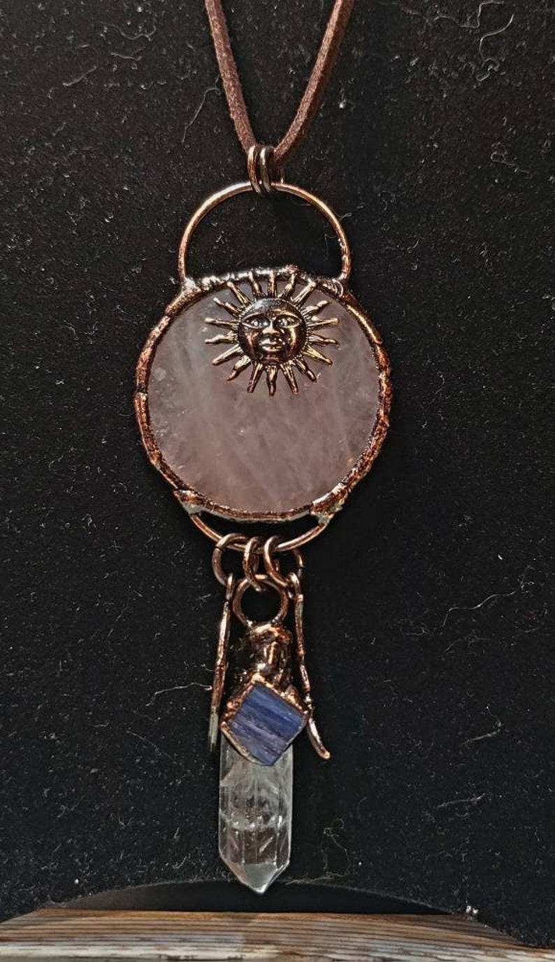 Op de afbeelding: Een ketting met een hanger van rozenkwarts, een koperen zonhanger en een punt van helder kwartskristal met een blauwe steenaanvulling. De ketting is aan een bruin leren koord.